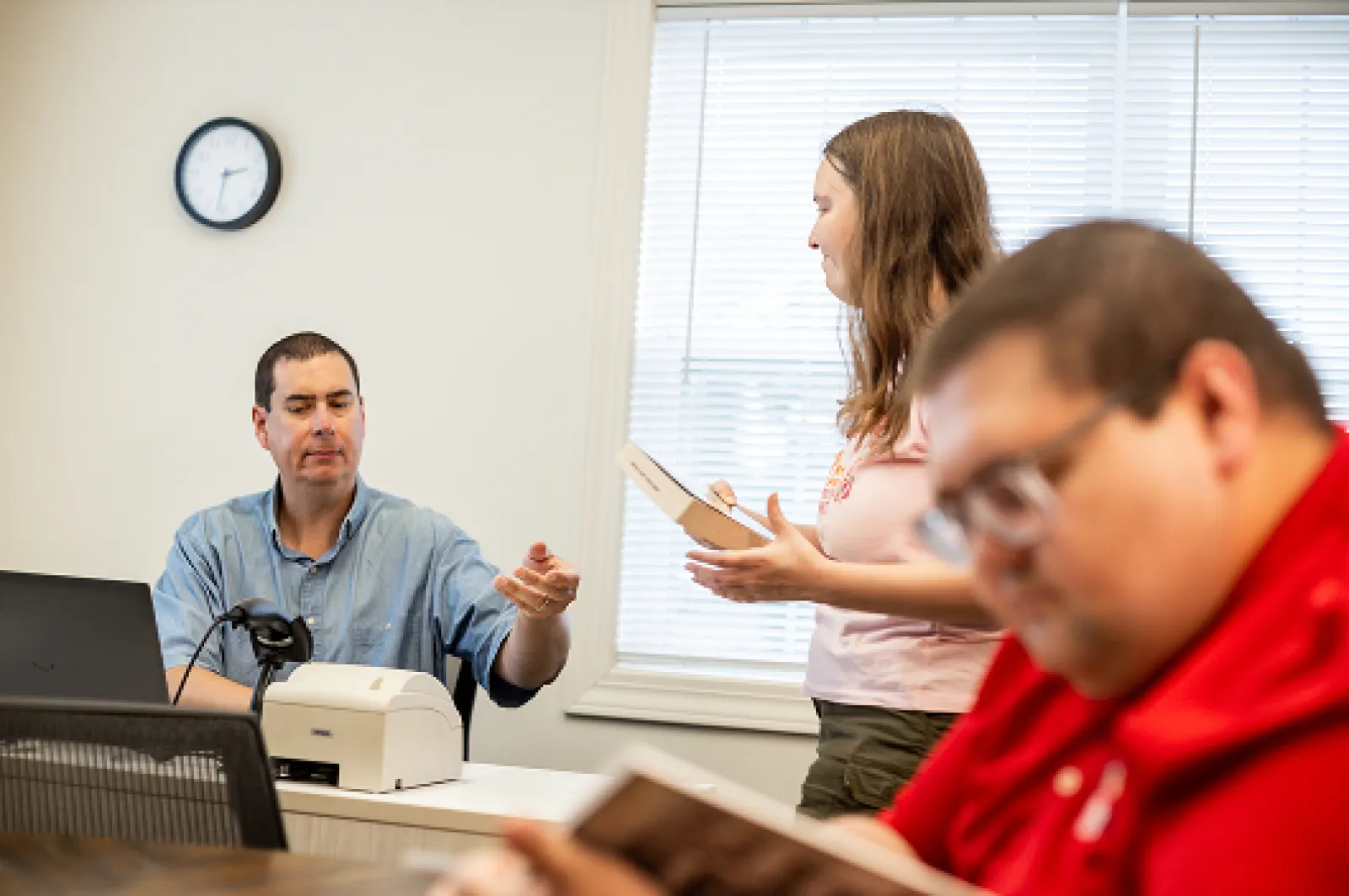 Female student handing a book to a librarian to checkout the book. A male student is reading in the foreground of the photo.