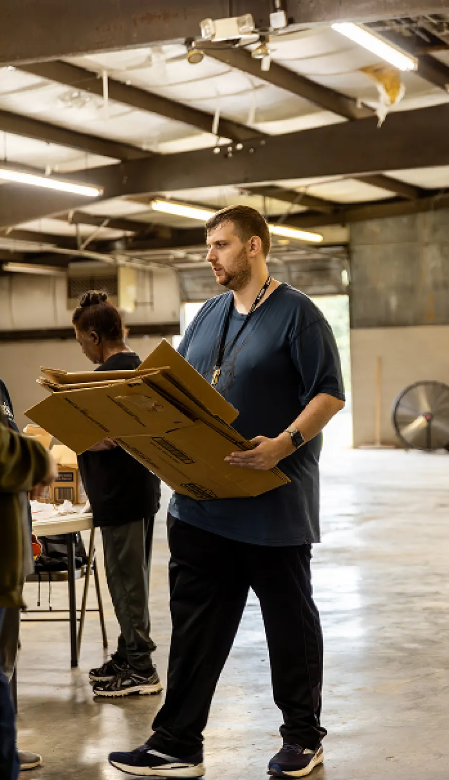 Male in a navy blue shirt is carrying cardboard boxes in a warehouse.